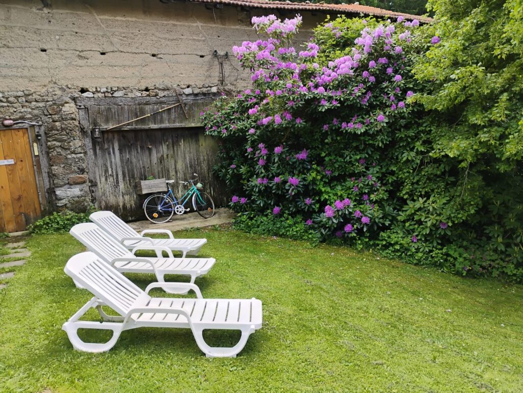 Jardin du Domaine de la Chouette avec transats, grange en bois et vélo ancien près d’un massif fleuri.