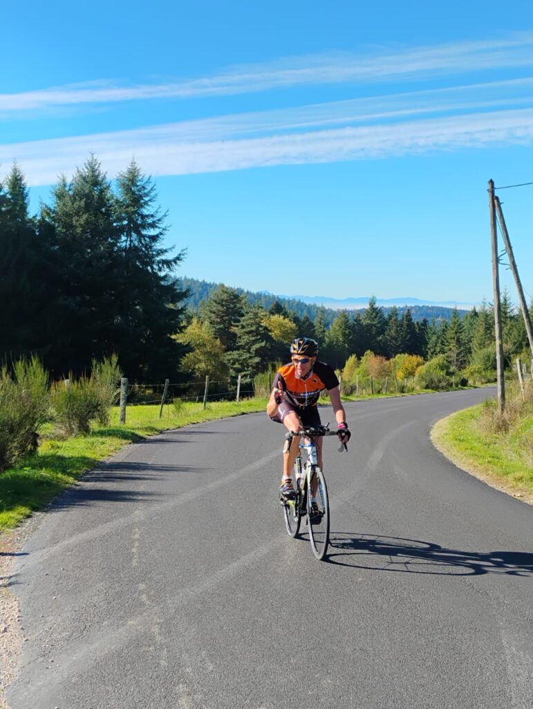 “Cycliste sur une route panoramique près du Domaine de la Chouette, entourée de sapins et de montagnes sous un ciel bleu.